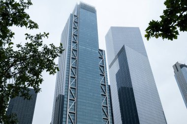 Yenilikçi geometrik tasarım kulesine sahip modern gökdelenler gür ağaç tepeleri üzerinde, dramatik bir bulutlu zemin karşısında kentsel büyümeyi sembolize ediyor.