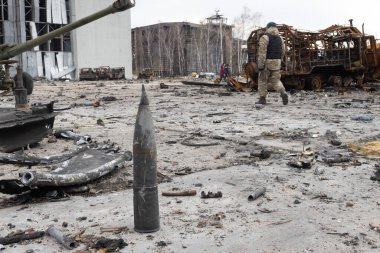 Savaşın parçaladığı Ukrayna 'da hırpalanmış bir obüs enkazın ortasında uzanıyor ve kamufle olmuş bir asker yakınlarda devriye geziyor. yıkılan bir bina ve yıkılan araçlar korkunç bir zemin boyuyor. 2022-04-05