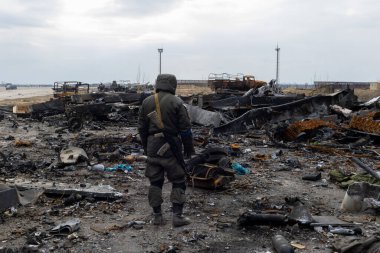 Savaşın parçaladığı Ukrayna manzarasında, mavi bir kol bandıyla tanımlanan bir asker, kamuflaj kıyafetleri içinde askeri araçların enkazını inceliyor. 2022-04-05