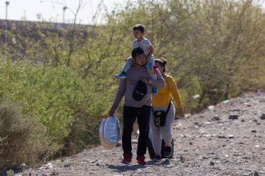 Bir aile Ciudad Juarez sınırında tozlu bir patika boyunca yürüyor, çocuk omuzlarında, göçün ortasında..