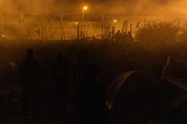 Ciudad Juarez 'deki aydınlık sınır çitleri ve devriye figürleriyle gece çadırların yakınındaki siluetli göçmenler..