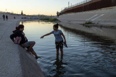 Gün batımında Ciudad Juarez 'deki bir sınır çitinin yanında, göçmenlik ve sınır geçişleri için önemli bir yer olan su kenarında sohbet eden iki kişi görülüyor..