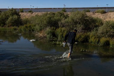 Bir göçmen, arka planda ABD sınır çiti olan Ciudad Juarez yakınlarındaki nehri geçiyor..