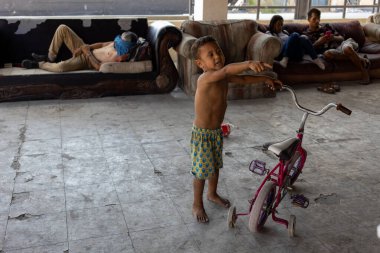 Genç bir çocuk, yıkık dökük bir odada ailesinin arasında bisikletle oynuyor. Ciudad Juarez sınırındaki göçmenler için zorlu yaşam koşullarını vurguluyor..