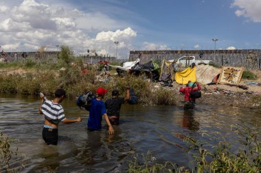 Göçmenler Ciudad Juarez yakınlarındaki bir nehirden geçtiler, açık gökyüzünün altında, çitli bir sınırın yakınındaki bir kampa ait eşyalar taşıyorlardı..