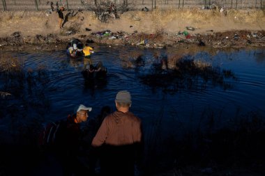 Göçmenler Ciudad Juarez yakınlarındaki enkaz dolu Rio Grande 'de yürürken, diğerleri düzensiz bir geçiş sırasında bankadan izliyorlar..