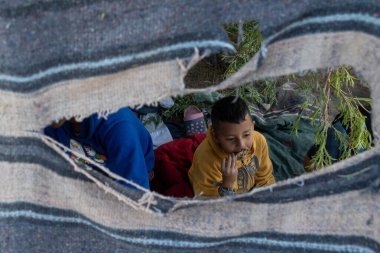 İki çocuk, Ciudad Juarez 'deki göçler sırasında yemek yerken yırtık kumaşın altına sığınıyor..