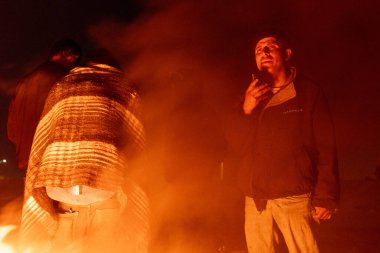 Erkekler, muhtemelen göçmenler, Ciudad Juarez yakınlarında bir ateşin başında toplanıp yolculuklarını tartışırlar..