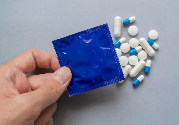 Yakın plan bir el, mavi bir prezervatif ambalajı içinde haplar ve kapsüller var.