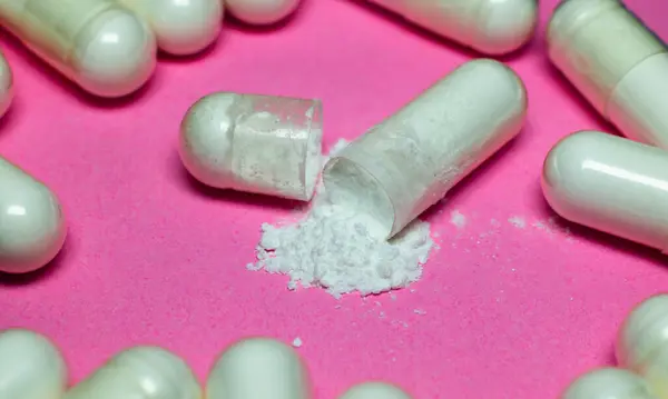 Kırık bir ilaç kapsülü daha fazla kapsülle çevrili pembe bir yüzeye beyaz toz saçıyor..