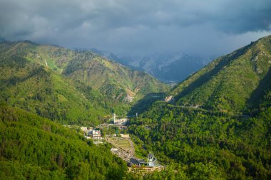 Yeşil orman yamaçlarıyla panoramik dağ vadisi manzarası ve fırtınalı gökyüzünün altında uzak karlı zirveler.
