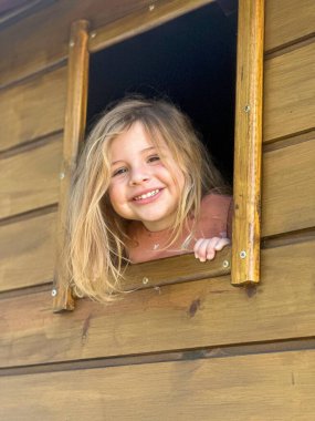 Menina loira sorridente aparecendo em uma janela de uma casa de madeira, exibindo uma express so feliz e acolhedora.