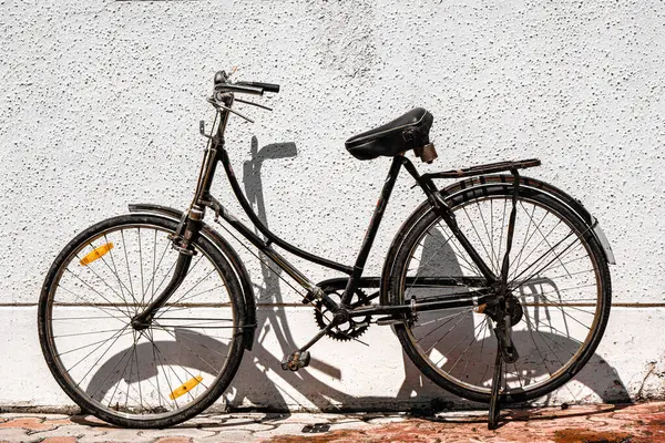 Eski, siyah eski bir bisiklet güneşli beyaz bir duvara dayanır, kaldırımda taze bir gölge bırakır günlük şehir hayatının nostaljik bir anı.