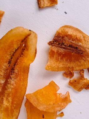 Düz beyaz bir arkaplanda sunulan üç kurutulmuş muz diliminin fotoğrafı. Dilimler, kurutma işleminden görülebilir dokusu olan zengin, altın-turuncu bir renk sergiliyor. Muzun içindeki doğal şekerler karamelize olmuş.