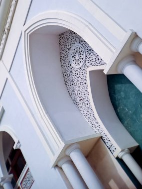 Karmaşık kemerli pencereleri ve balkonları olan beyaz cami cephesi açık mavi gökyüzüne doğru uzanıyor.