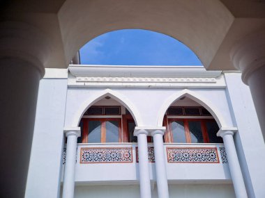 Kemerli camları ve balkonları olan beyaz bir cami cephesinin çerçeveli görüntüsü, canlı mavi gökyüzüne karşı kurulmuş.