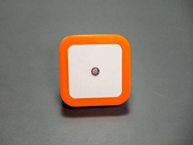 Yuvarlak kare forma sahip bir geometrik nesne, beyaz, turuncu ve koyu gri arkaplan etkileşimini vurguluyor, nötr zemin üzerinde modern basitliği sergiliyor..