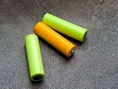 Üç silindirik nesne, iki canlı yeşil ve bir parlak turuncu, basit, süslenmemiş bir stüdyo düzeninde koyu gri bir yüzey üzerinde dinleniyor, temiz ve minimal bir kompozisyon yaratıyor..