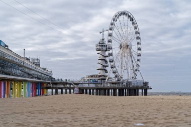 Scheveningen, Hollanda - 30 Mart 2025: Bulutlu bir günde dönme dolabı ve plajı olan Total Pier, geniş açıyla sahilde oturan kadın