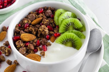Greek yogurt with chocolate granola, kiwi fruit, pomegranate seeds and almonds in a bowl for healthy breakfast on white background. Close up