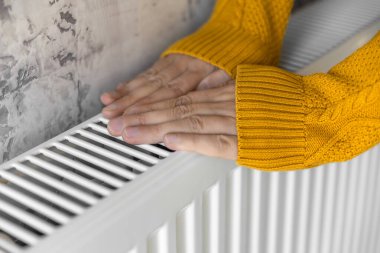 Closeup of man warming his hands in yellow sweater on the heater at home during cold winter days. Male getting warm up his arms over radiator. Concept of heating season or cold weather