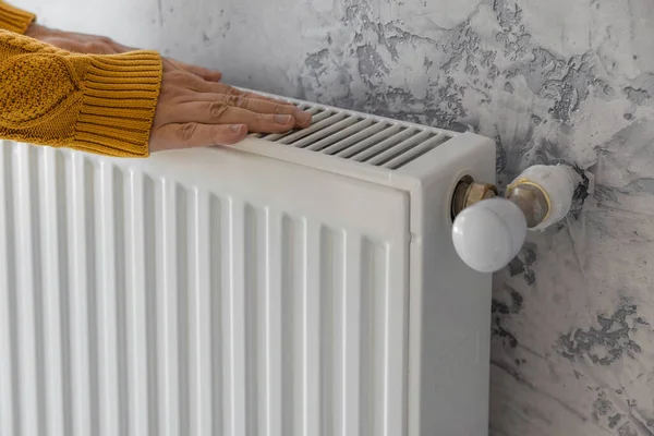 Man in yellow sweater warming his hands on the heater at home during cold winter days. Male getting warm up his arms over radiator. Concept of heating season or cold weather