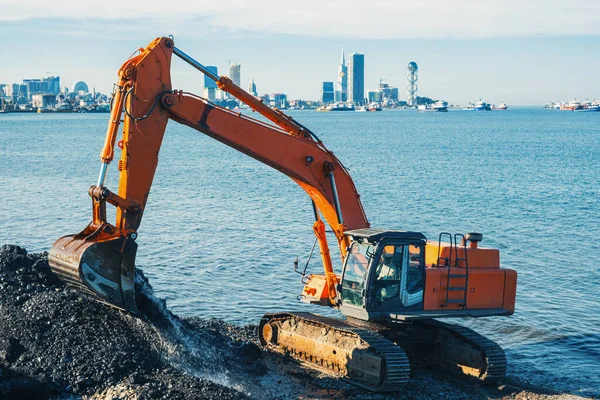 Excavator or digger working on earthmoving at shore protection works in Batumi. Orange backhoe digs sand and gravel in quarry. Digger during excavation