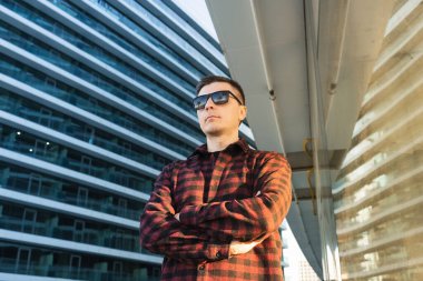 Olumlu, neşeli, güvenilir bir serbest çalışan portresi ya da çapraz kollu bilişim uzmanı. Güneş gözlüklü bir adam modern ofis binalarının önünde duruyor. Güneş gözlüklü bir plaj yansıması var. Komik atış.