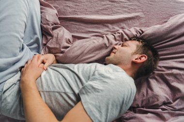 Hüsrana uğramış yakışıklı genç adam evde kötü bir şekilde yatarken karnına sarılıyor. Üst Manzara. Karnı ağrıyan bir erkek. Mide ülseri, mide iltihabı, gıda zehirlenmesi.