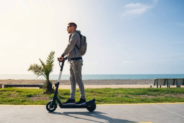 Genç bir gezgin, güneşli yaz gününde deniz kıyısı ve plaj boyunca e-scooter kullanıyor. Erkek, dışarıda elektrikli scooter kullanıyor. Sürdürülebilir taşıma ve seyahat. Sürdürülebilir yaşam tarzı kavramı