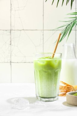Beyaz fayans zemininde, sert gölgeler ve palmiye yapraklarıyla kaplı bir bardakta buzlu yeşil kibrit latte. Soğuk yaz içkisi. Dikey yönelim