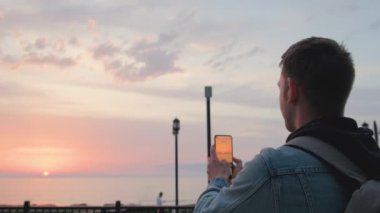 Gezgin adam cep telefonundaki kamerayla deniz batımını fotoğraflıyor. Beyaz adam deniz kıyısında beklerken akıllı telefondan fotoğraf çekiyor. Yaz tatili konsepti