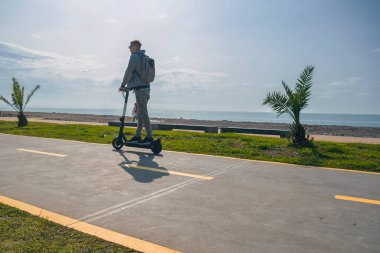 Genç bir gezgin, güneşli yaz gününde deniz kıyısı ve plaj boyunca e-scooter kullanıyor. Erkek, dışarıda elektrikli scooter kullanıyor. Sürdürülebilir taşıma ve seyahat. Sürdürülebilir yaşam tarzı kavramı