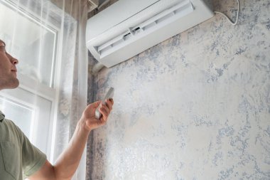Genç bir kadın sıcak yaz mevsiminde konfor sıcaklığı için evdeki klimayı uzaktan kumandayla açıyor. Modern ev teknolojisi