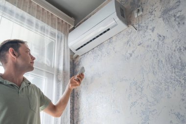 Genç bir kadın sıcak yaz mevsiminde konfor sıcaklığı için evdeki klimayı uzaktan kumandayla açıyor. Modern ev teknolojisi