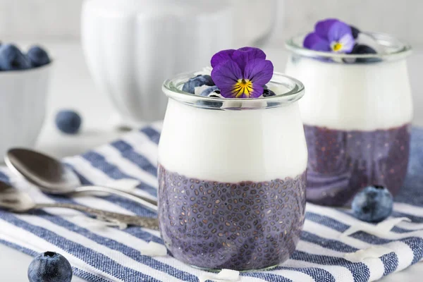 stock image Healthy vegan breakfast of blueberry yogurt parfaits made with fresh berries, coconut yoghurt, chia pudding and edible flowers in glasses over napkin on white table