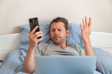 Cep telefonuyla tartışan sinirli bir adam evde bir yatakta yatıp bilgisayar sorununu akıllı telefonuyla çözerken dizüstü bilgisayara bakıyor. Kablosuz teknoloji