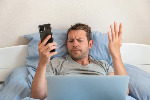 Rozzlobený Frustrovaný Muž Mobilním Telefonem Při Pohledu Notebook Při Ležení — Stock fotografie