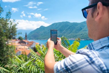 Güneş gözlüklü adam Karadağ 'ın Kotor Körfezi' ndeki eski Perast kasabasının fotoğrafını çekiyor. Yaz tatilindeki erkek turist sosyal medya için dönüm noktası fotoğrafları çekiyor. Genç adam tatilin tadını çıkarıyor. Seyahat ve