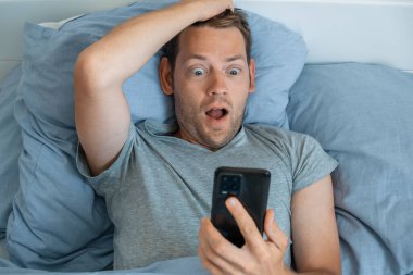 Akıllı telefondan haber okuyan yakışıklı bir adam sabah yatakta şok olmuş bir ifadeyle. Adam cep telefonundan alınan haberlere hayran, elinde bir telefon, ağzıyla ekrana bakıyor.