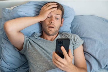 Akıllı telefondan haberleri okuyan yakışıklı bir adam. Sabahları yatakta üzgün bir ifadeyle. Adam cep telefonundan gelen kötü haberlere hayret ediyor, telefonu tutuyor ve ağzıyla ekrana bakıyor.