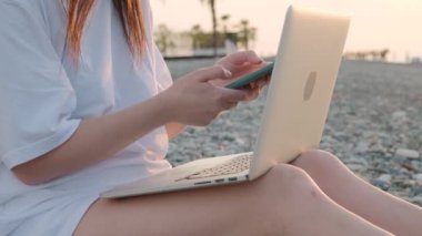 Gün batımında sahilde dizüstü bilgisayarı ve cep telefonuyla çalışan tanınmayan bir kadına yakın çekim. Serbest çalışan bir kadın uzaktan akıllı telefon ve bilgisayar kullanarak çevrimiçi çalışıyor. Uzaktan çalışma