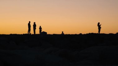 Deniz kıyısında siluetler oluşturan insanlar gün batımında güneşin batışını izliyorlar. Aile akşamı dışarıda..