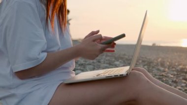 Gün batımında sahilde dizüstü bilgisayarı ve cep telefonuyla çalışan tanınmayan bir kadına yakın çekim. Serbest çalışan bir kadın uzaktan akıllı telefon ve bilgisayar kullanarak çevrimiçi çalışıyor. Uzaktan çalışma
