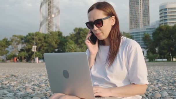 数字游牧民族快乐的女人带着笔记本电脑在海边用手机聊天 女自由职业者通过智能手机和远程计算机在网上工作 远程工作 — 图库视频影像