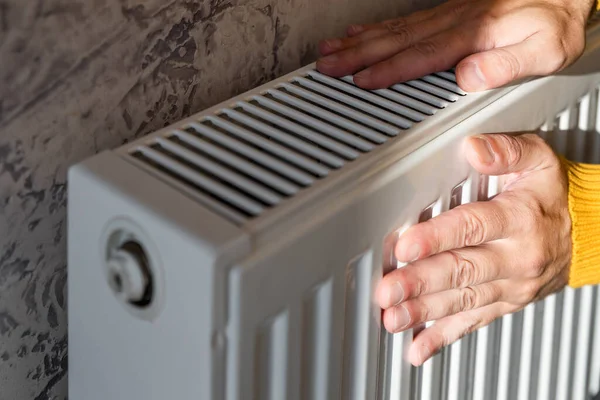 Man warms his hands on the radiator at home on cold sunny winter day. Male getting warm up his arms over heater. Concept of heating season or cold weather.