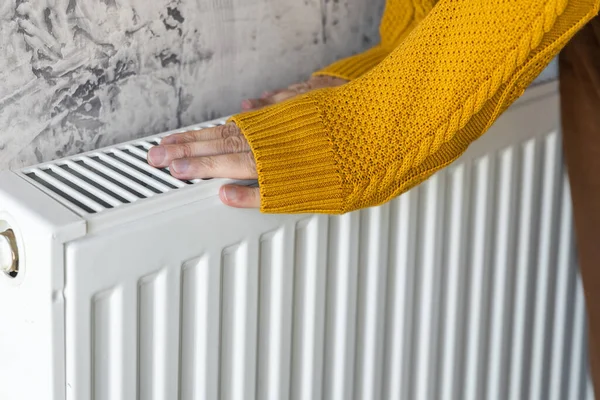 Man warming his hands in yellow sweater on the heater at home during cold winter days. Male getting warm up his arms over radiator. Concept of heating season or cold weather.