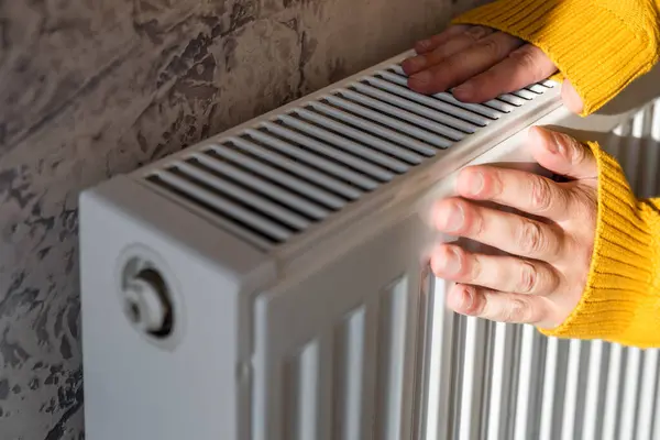Man warms his hands on the radiator at home on cold sunny winter day. Male getting warm up his arms over heater. Concept of heating season or cold weather.