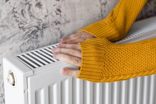 Close up of man warming his hands in yellow sweater on the heater at home during cold winter days. Male getting warm up his arms over radiator. Concept of heating season or cold weather.