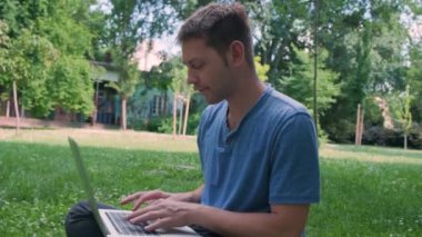 Mavi tişörtlü genç serbest çalışan adam yeşil çimlerin üzerinde oturur ve doğada dizüstü bilgisayarla çalışır. Erkek öğrenci dışarıda bilgisayarda daktilo kullanıyor. Uzak meslek, çevrimiçi eğitim kavramı.